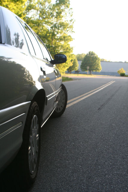 2004 Ford Crown Victoria - A silver sedan is parked on the side of a road with trees and a building in the background.