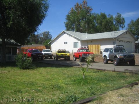 1990 Ford Bronco - Several lifted off-road trucks, including a red prerunner and white Bronco, parked in the driveway of a single-story house.