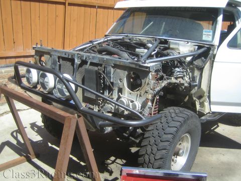 1990 Ford Bronco - white pickup with its front clip removed sits on a steel stand, exposing the bare frame, a black tube bumper and a row of round gauges.