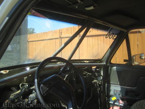 1990 Ford Bronco - Inside a stripped cab a roll cage runs across the windshield, with a Ford-emblem steering wheel and dusty glass overlooking a wooden fence.