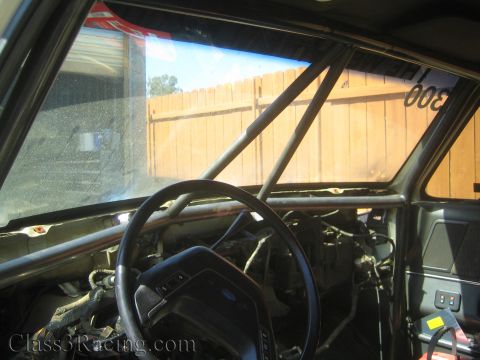 1990 Ford Bronco - View across a gutted cab interior with a roll cage bar, a Ford steering wheel and a stripped dash, framed by the windshield and a wooden fe…