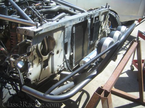 1990 Ford Bronco - Side view of a project truck's bare front frame on a stand, with tube bumper, mounted round light housings and exposed radiator support.