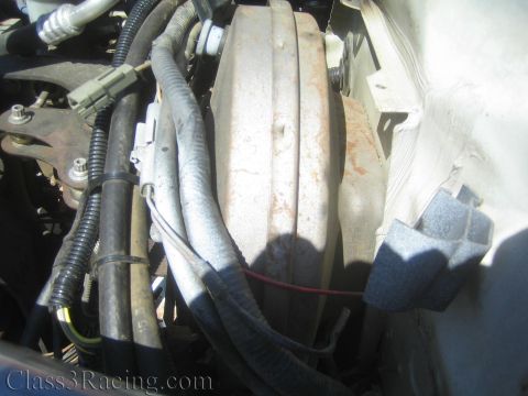 1990 Ford Bronco - Engine-bay detail showing a large round metal canister surrounded by loomed wiring, hoses and gray foam padding against a white body panel.