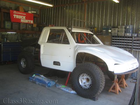 1990 Ford Bronco - white fiberglass-bodied off-road race truck sits on jack stands in a metal shop, beneath a red Dodge Truck Month banner.