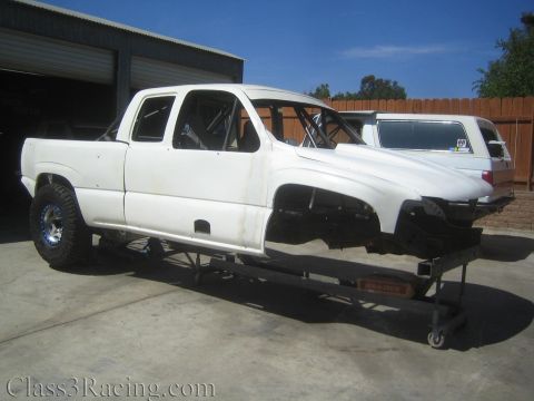 1990 Ford Bronco - white extended-cab truck body sits on a low rolling cart outdoors, missing its front clip, with a covered camper-topped truck behind it.