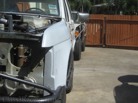 1990 Ford Bronco - white front fender and partly stripped front clip of a lifted truck, with an amber side marker, parked on concrete by a red wood fence.