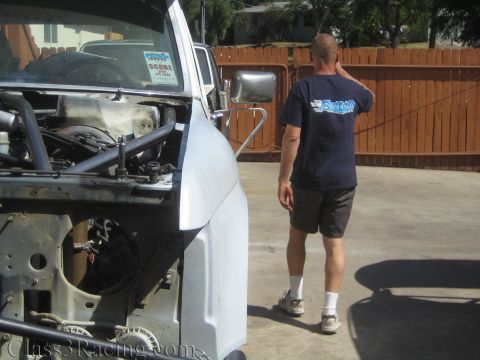 1990 Ford Bronco - man in a dark T-shirt and shorts walks across a concrete yard past a white truck whose front clip and engine bay are exposed.