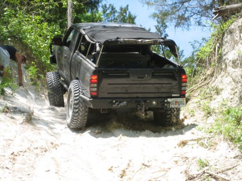 1989 Ford Bronco - A modified black pickup truck navigates a sandy off-road path with dense foliage on the sides.