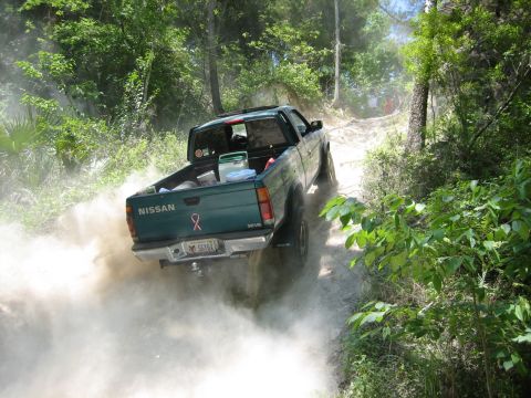 1989 Ford Bronco - A green Nissan pickup truck navigates a dusty off-road trail, kicking up a cloud of dirt.