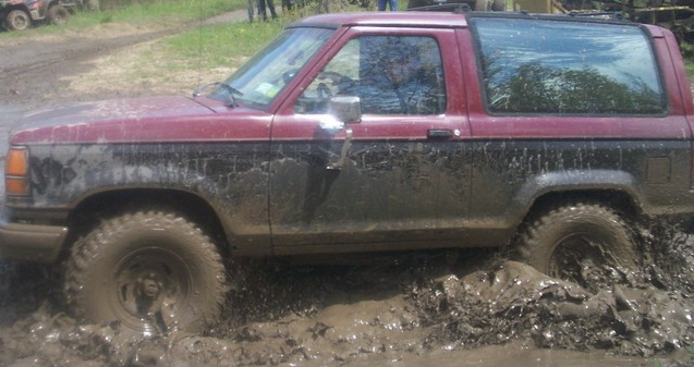 Red and black Ford Bronco II struggling in deep mud, front wheel submerged