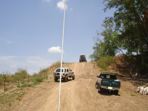 1989 Ford Bronco - Several off-road vehicles ascend a challenging dirt incline under a clear blue sky.