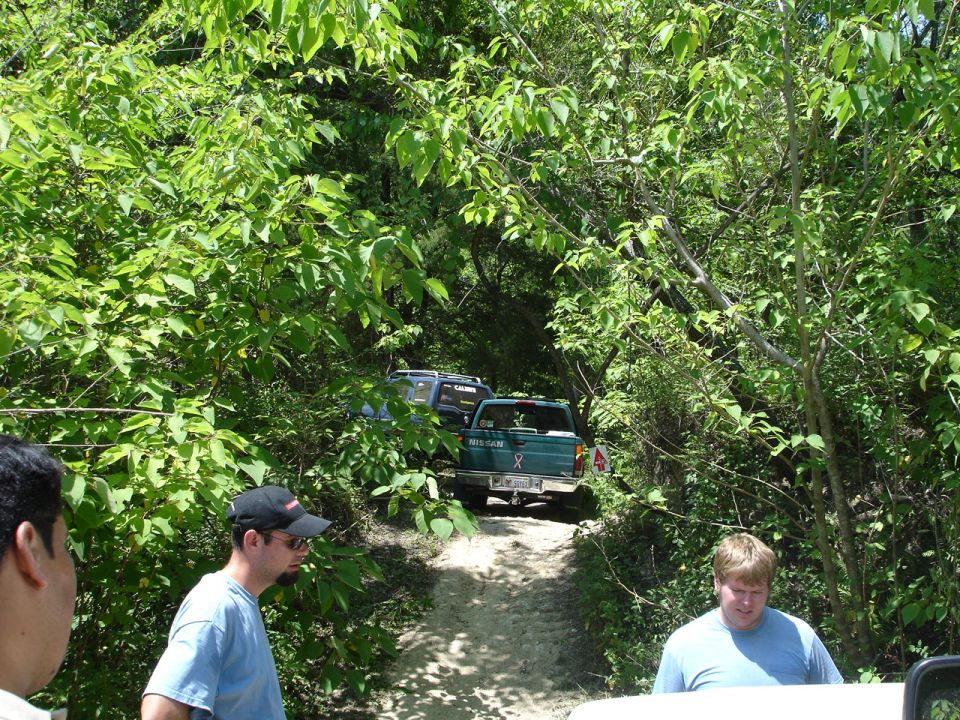 Two trucks driving on a dirt path through trees. People are visible in the foreground