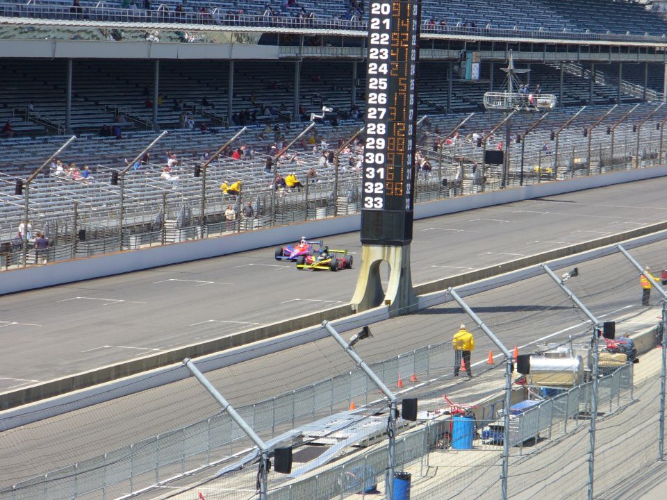 Two IndyCars on a race track with a scoring tower displaying numbers 20-33