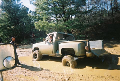 1989 Ford Bronco - A pickup truck is stuck in a muddy off-road area with people standing nearby.