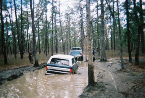 1989 Ford Bronco - Two SUVs navigate a waterlogged forest path, with one vehicle partially submerged.