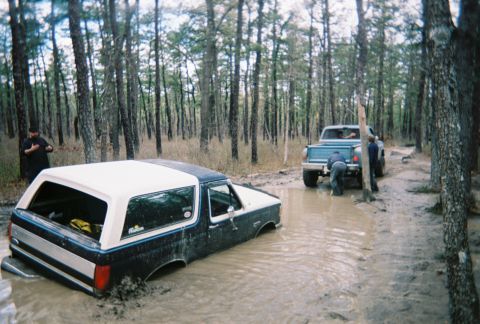 1989 Ford Bronco - Two vehicles are stuck in a muddy forest path, with people trying to free them.