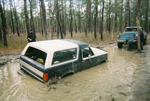 1989 Ford Bronco - A 1987 Ford Bronco is deeply stuck in a muddy clearing, while a teal pickup truck is also in the mud nearby.