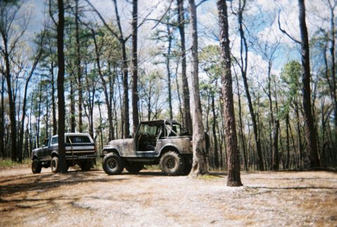 1989 Ford Bronco - Two off-road vehicles are parked in a forest clearing under a partly cloudy sky.