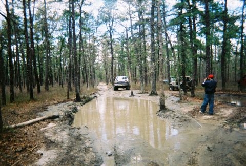 1989 Ford Bronco - A white 1989 Ford Bronco navigates a water-filled, muddy path in a wooded area, with another vehicle visible in the background.