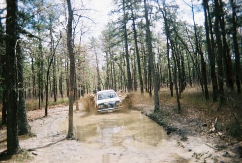 1989 Ford Bronco - A 1989 Ford Bronco navigates a challenging muddy path surrounded by trees.