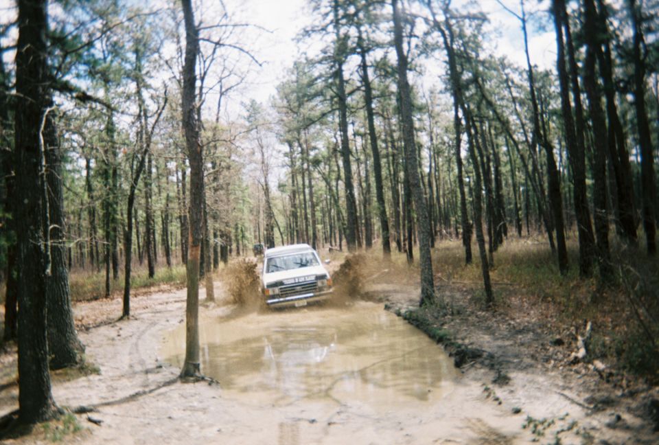 1989 Ford Bronco driving through a muddy trail in a wooded area