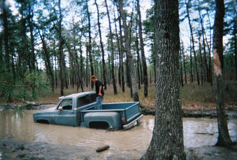 1989 Ford Bronco - A man stands in the bed of a blue pickup truck that is submerged in mud within a wooded area.