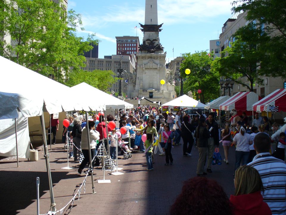 Outdoor festival with tents, crowds, and a tall monument in the background