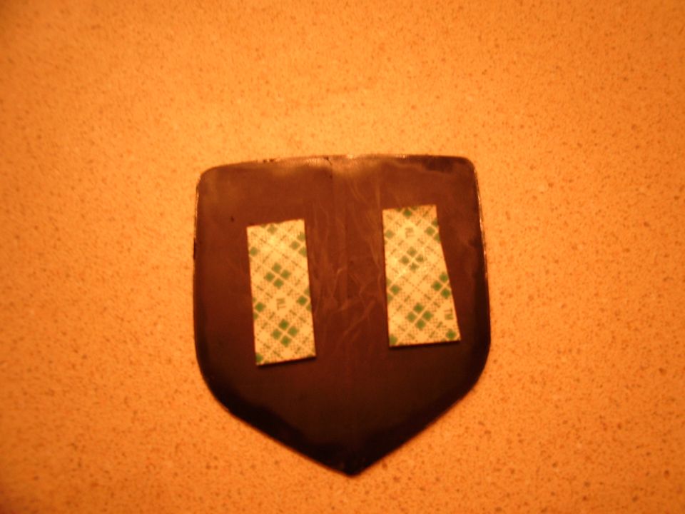 dark shield-shaped emblem with two strips of green and white adhesive tape on the back