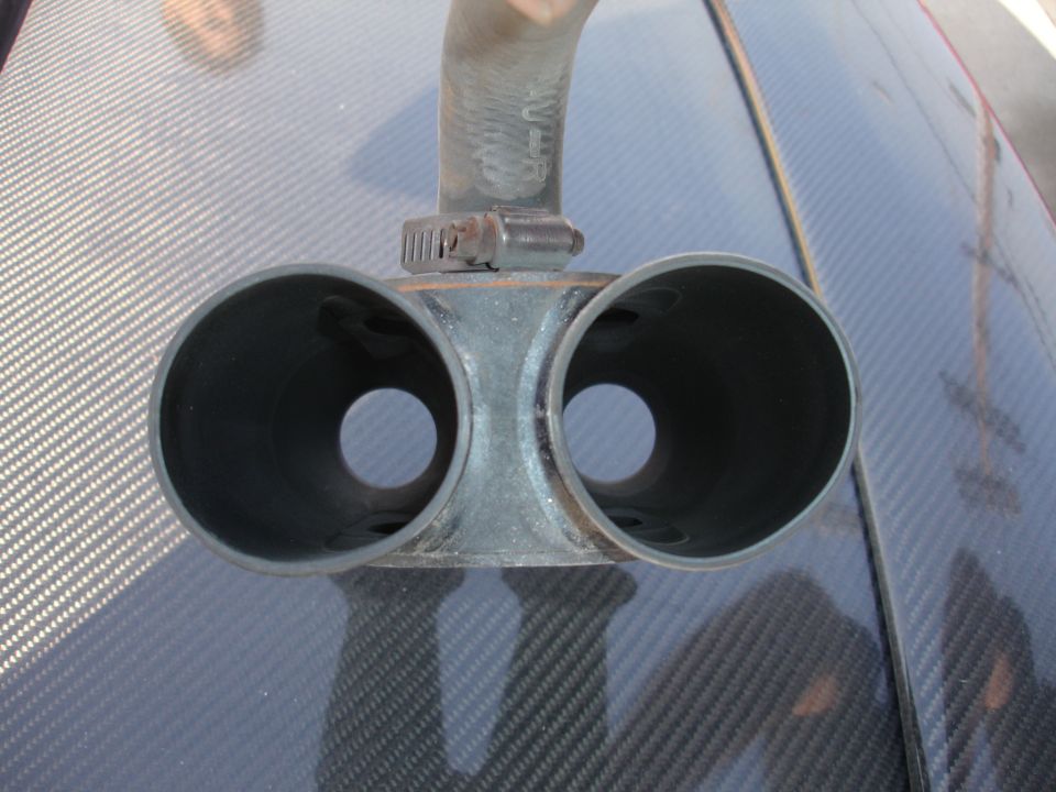 Dual exhaust tip with a hose clamp and rubber hose attached, resting on a carbon fiber surface
