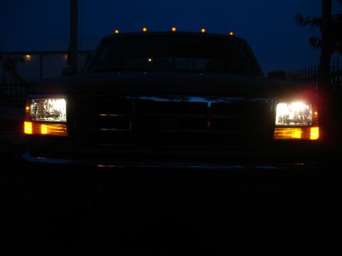 1992 Ford F-350 - The front of a Ford F-350 truck is illuminated by its headlights and roof marker lights against a dark blue sky.