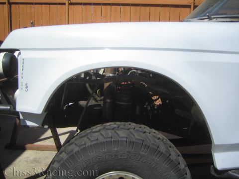 1990 Ford Bronco - wide white front fender flare on a lifted truck arches over a BFGoodrich tire, with airbag suspension visible in the wheel well by a wood f…