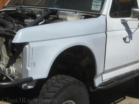1990 Ford Bronco - white front fender and cab of a lifted truck with its hood raised, side step bar below, parked on a concrete pad outdoors.