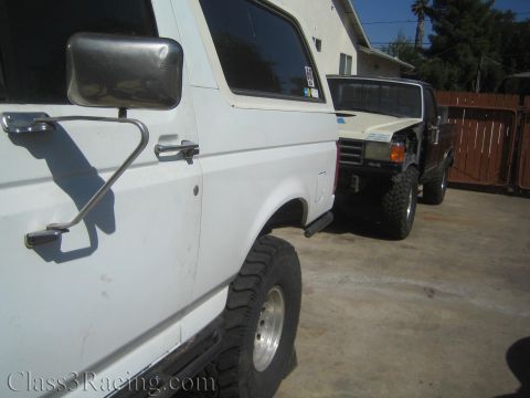 1990 Ford Bronco - white SUV with a chrome tubular mirror parked on concrete beside a darker lifted SUV with off-road tires, near a wood fence.