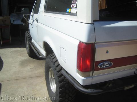 1990 Ford Bronco - Rear quarter of a white lifted SUV with two-tone lower trim, oval taillight and a Ford-badged tailgate, sitting on a mud-terrain tire.