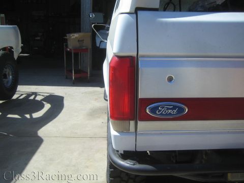 1990 Ford Bronco - Straight-on rear view of a white lifted Ford SUV showing the oval taillight and blue Ford oval badge on its two-tone tailgate in a shop.