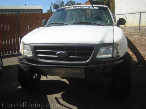 1990 Ford Bronco - Front view of a white lifted Ford pickup with a black tube bumper, Ford oval in the grille and a windshield banner, parked by a brick wall.