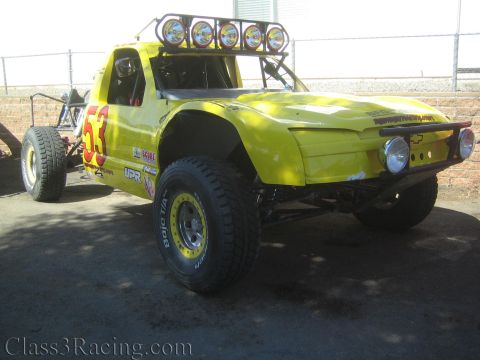1990 Ford Bronco - yellow off-road race truck marked 53 sits in a fenced lot, fitted with a five-light overhead bar, tube chassis and off-road tires.