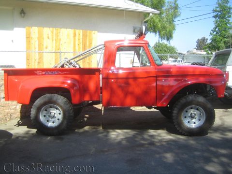 1990 Ford Bronco - Side profile of a red lifted Ford stepside pickup on chrome wheels and large tires, parked on a driveway by a wood fence and house.