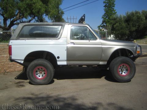 1990 Ford Bronco - lifted two-tone silver-and-white full-size two-door SUV sits in profile on a dirt driveway with chrome wheels.