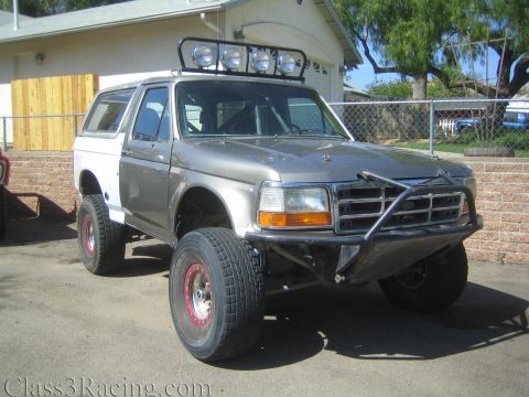 1990 Ford Bronco - gray lifted Ford SUV with a camper shell, roof-mounted light bar, and tubular prerunner front bumper parked by a house.
