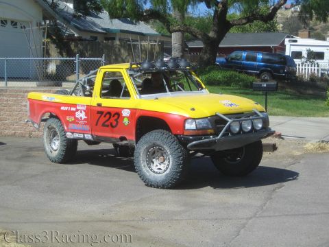 1990 Ford Bronco - yellow-and-red off-road race truck marked 723 with roof spotlights and a tubular bumper parked in a driveway.