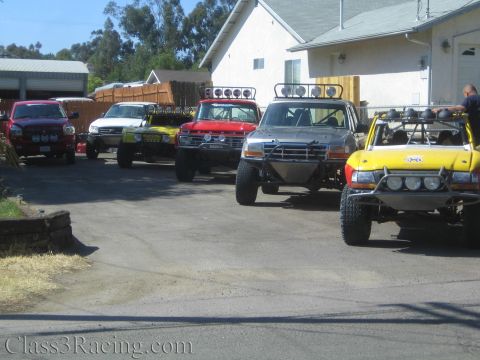 1990 Ford Bronco - Several off-road prerunner trucks and buggies, including red and yellow ones, parked in a row in a residential driveway.