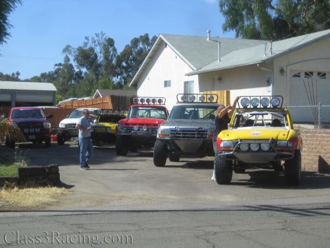 1990 Ford Bronco - lineup of lifted off-road prerunner trucks parked outside a house, with a man standing next to one of them.