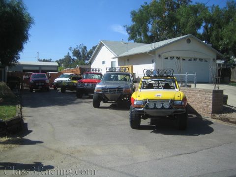 1990 Ford Bronco - group of lifted off-road prerunner trucks, including a yellow one in front, parked along a driveway by a house.