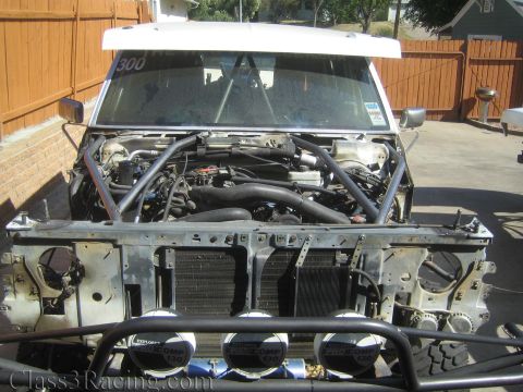 1990 Ford Bronco - stripped race truck front end showing the engine and roll cage, with three Pro Comp off-road lights mounted below.