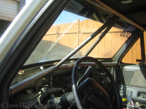 1990 Ford Bronco - Looking into a gutted truck cab showing a steering wheel, roll cage bars, and a wooden fence visible through the windows.