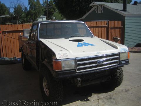 1990 Ford Bronco - white lifted Ford pickup with a blue arrow logo on the hood and 4x4 badging, parked on a concrete pad by a fence.