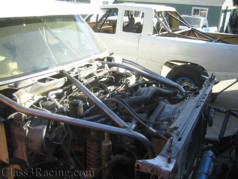 1990 Ford Bronco - exposed engine bay of a stripped race truck with a white prerunner truck parked behind it in a driveway.
