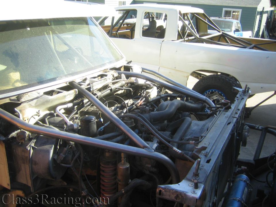Open engine bay of a stripped truck beside a white truck in a driveway