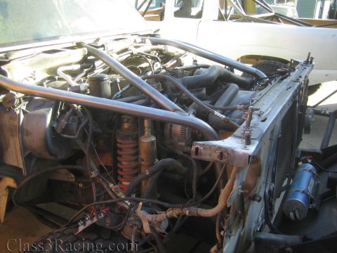1990 Ford Bronco - close-up of a race truck engine bay showing a coilover shock, alternator, roll cage tubing, and wiring harnesses.